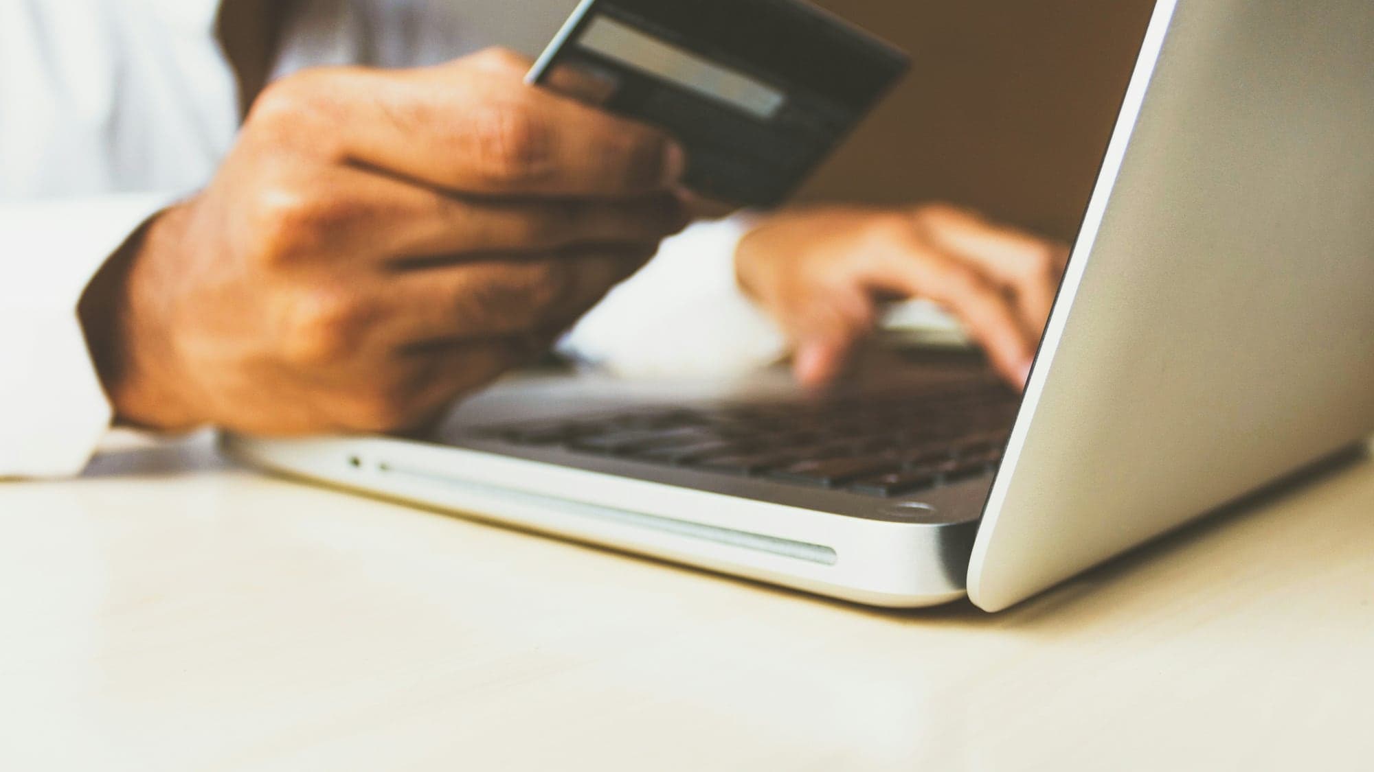 Mains d'un homme devant un ordinateur portable ouvert, une carte bancaire dans une main, l'autre sur le clavier, au moment de payer en ligne