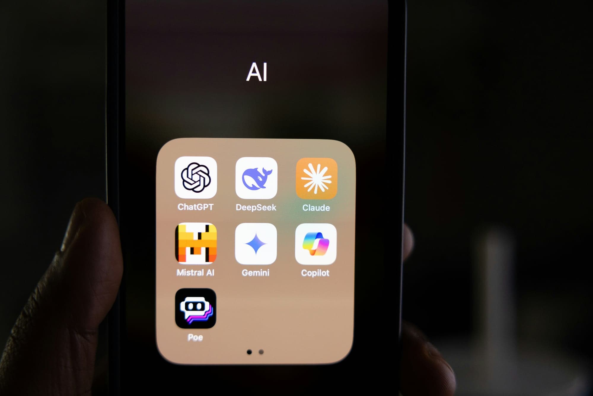 Main tenant un smartphone dont l'écran affiche un dossier « AI » regroupant les applications ChatGPT, DeepSeek, Claude, Mistral AI, Gemini, Copilot et Poe