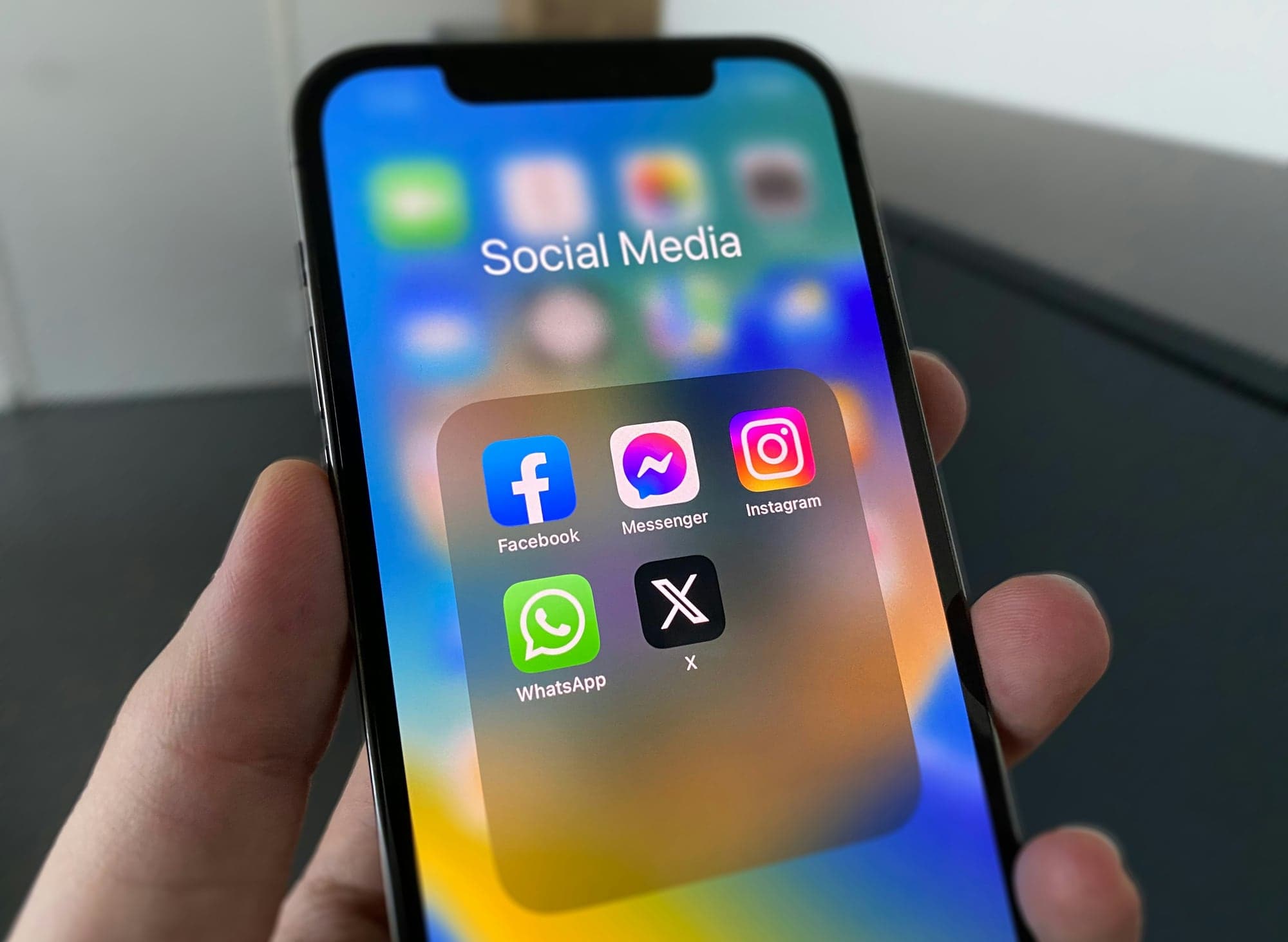 Une main tient un smartphone dont l'écran d'accueil affiche un dossier « Social Media » regroupant les applications Facebook, Messenger, Instagram, WhatsApp et X