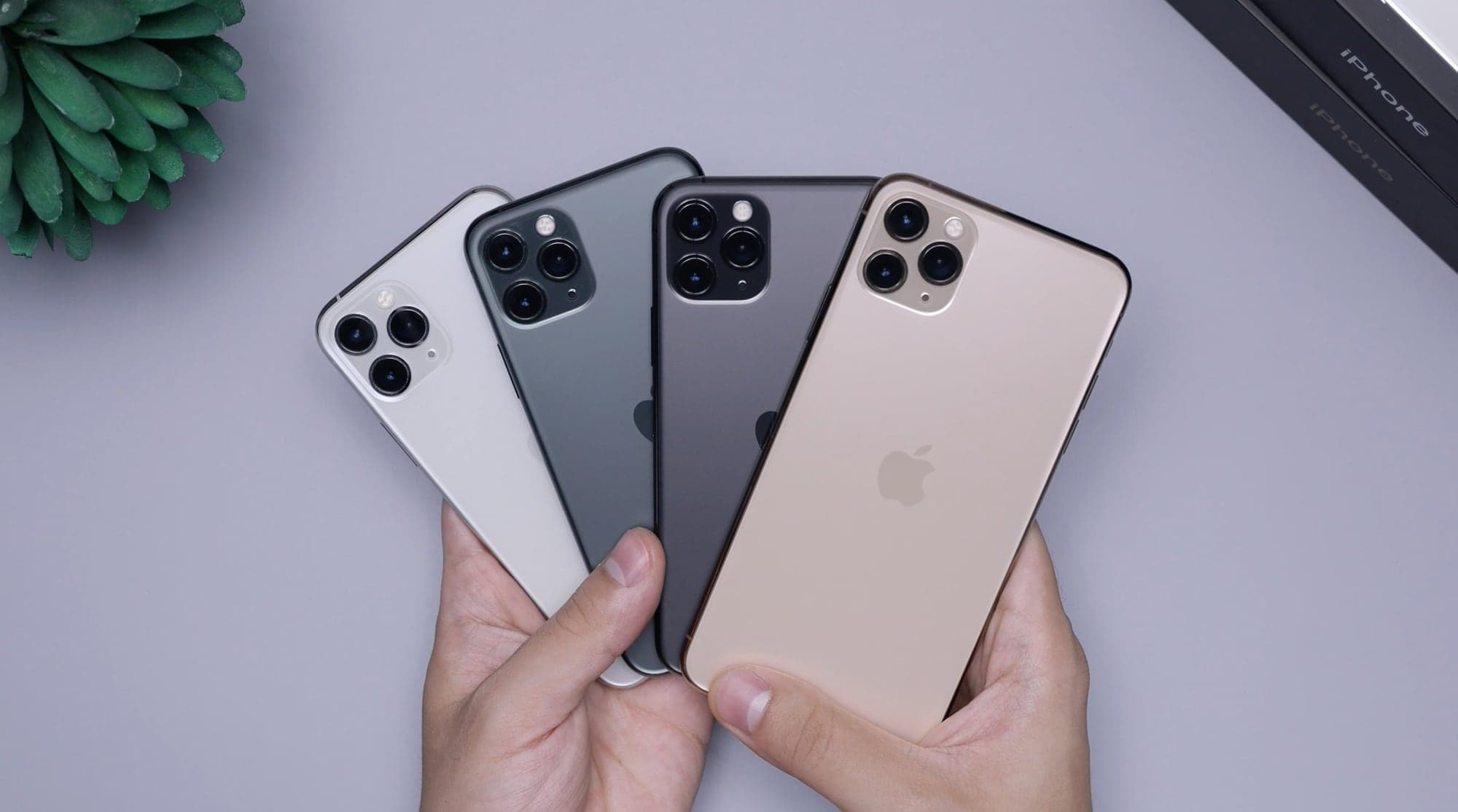 Deux mains tiennent en éventail quatre iPhone de la gamme 11 Pro, vus de dos, en argent, vert nuit, gris sidéral et or