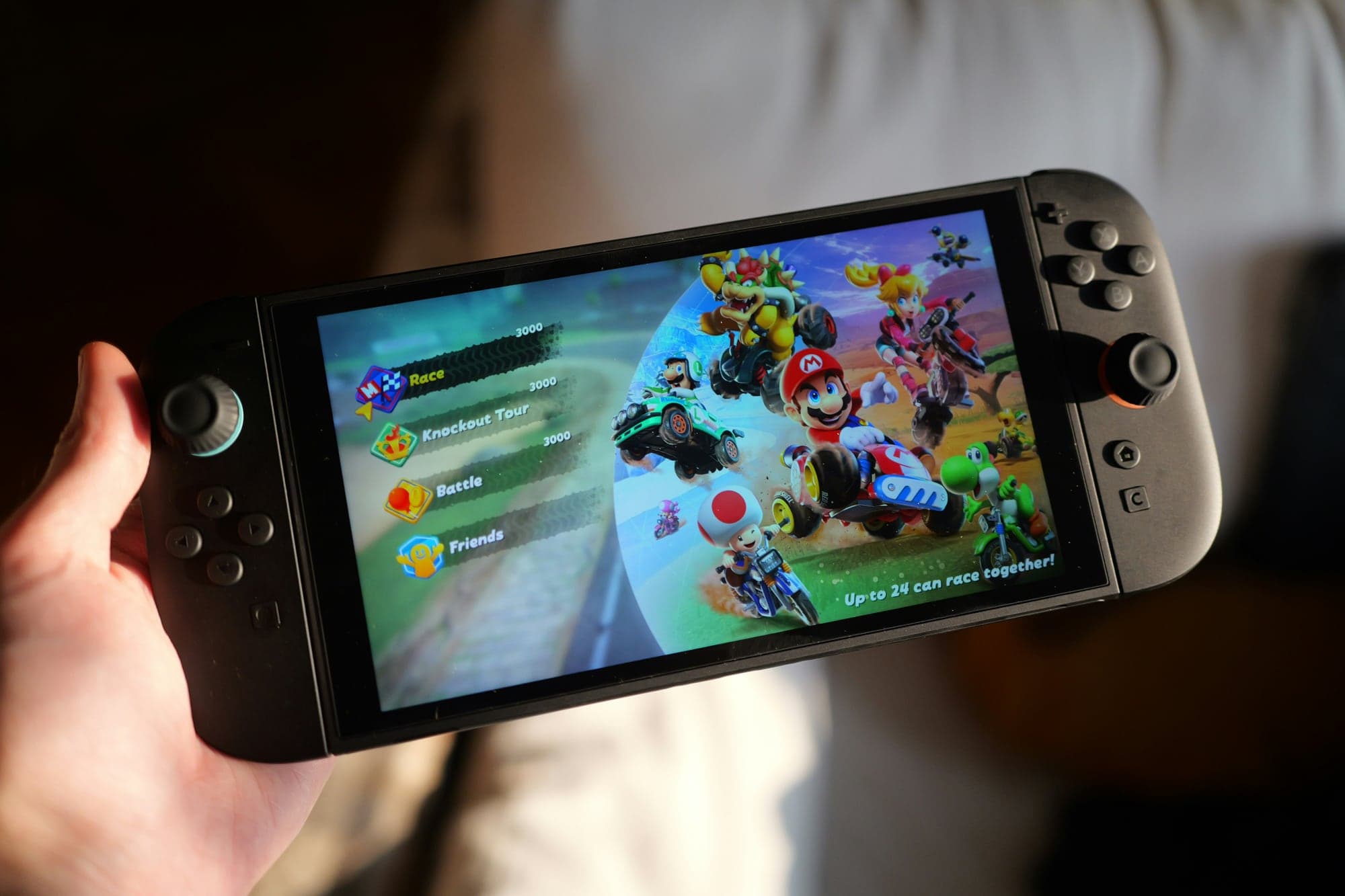 Une main tient une Nintendo Switch 2 en mode portable ; l'écran affiche le menu de Mario Kart World (Race, Knockout Tour, Battle, Friends) à côté d'une illustration de Mario, Luigi, Bowser, Toad et Yoshi en kart et à moto.