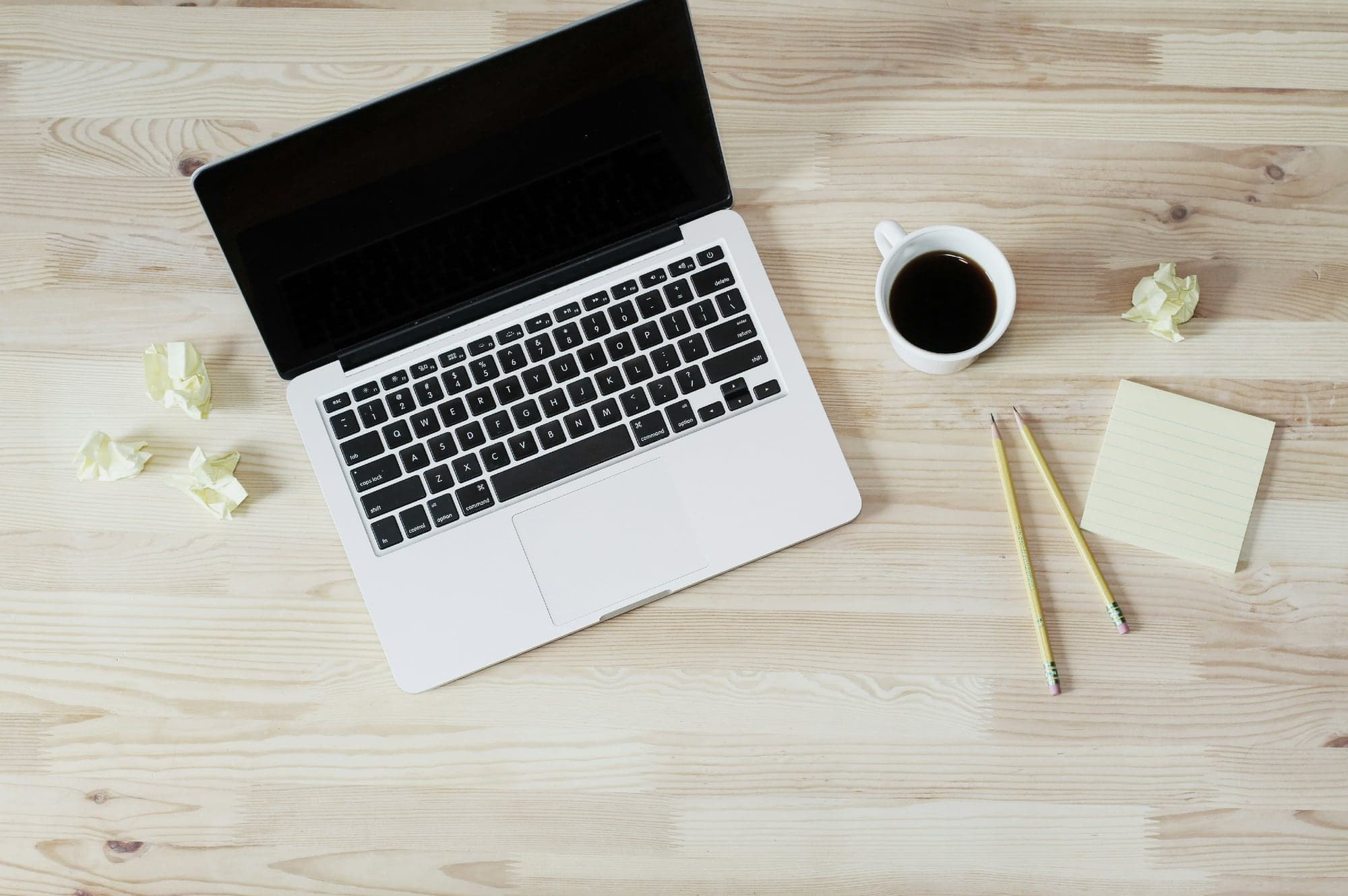 Vue de dessus d'un bureau en bois clair : un ordinateur portable à l'écran éteint, une tasse de café, des boulettes de papier froissé, deux crayons et un bloc-notes vierge