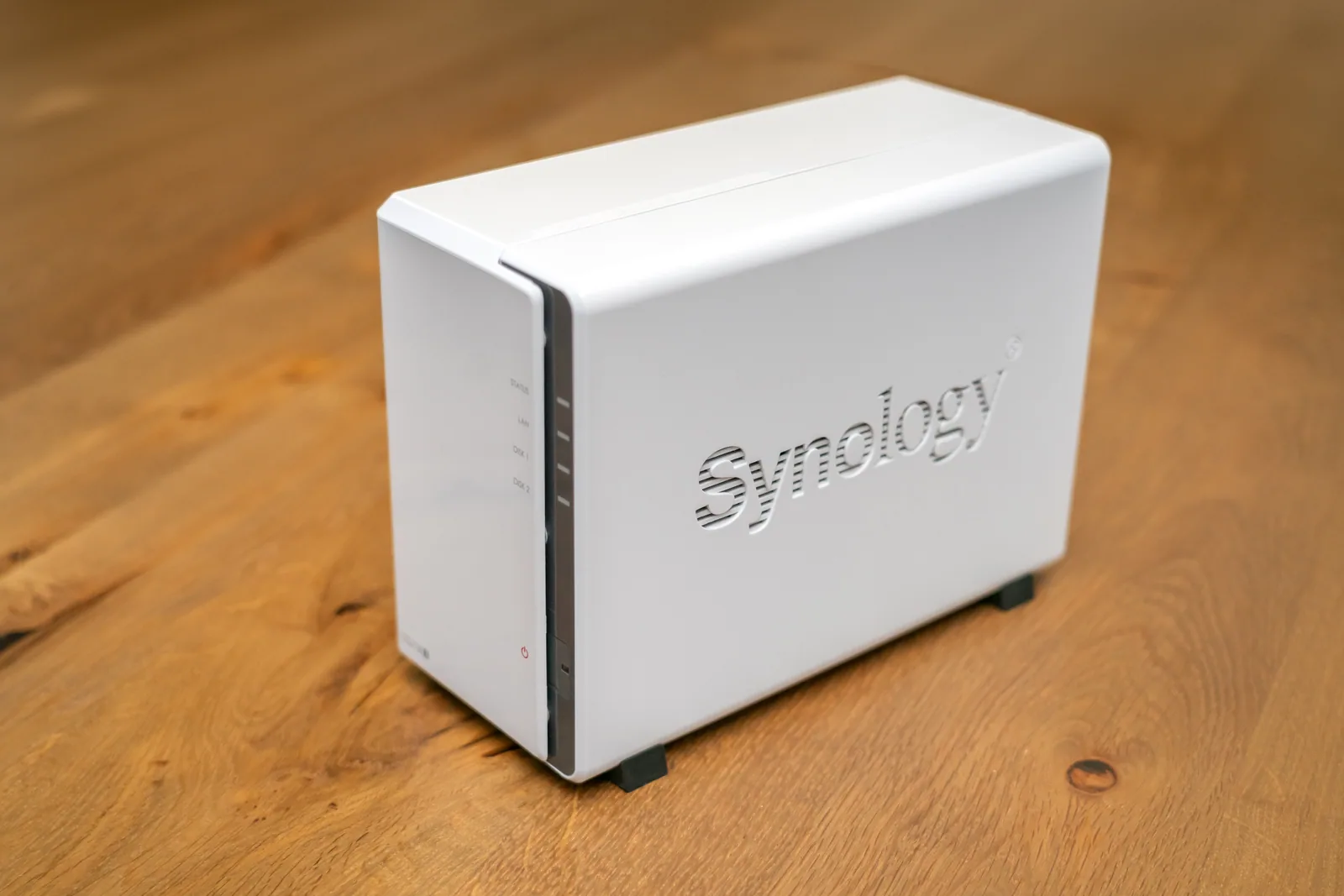 NAS Synology à deux baies et boîtier blanc, posé sur une table en bois, avec sur sa façade les voyants d'état, de réseau et des deux disques