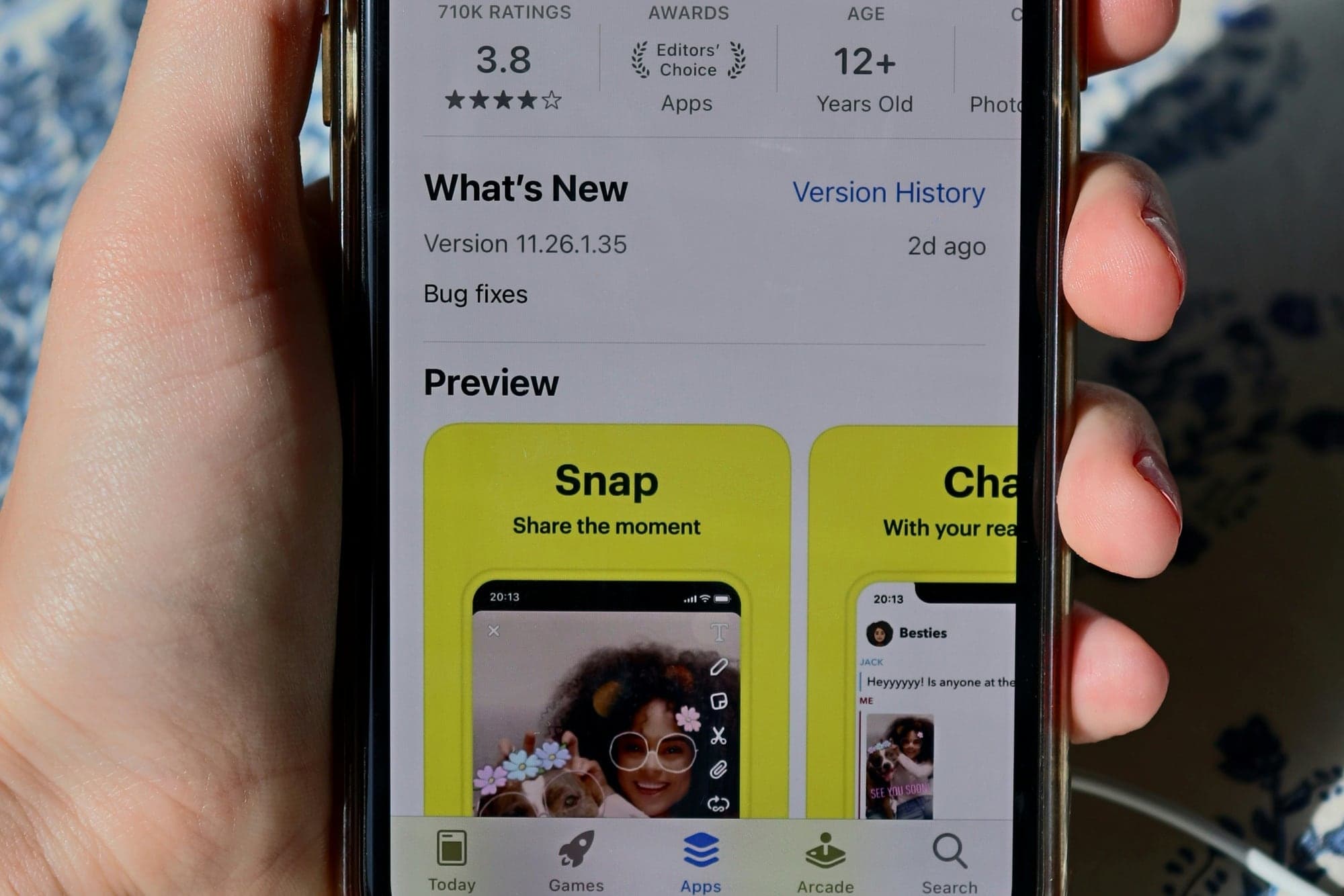 Main tenant un iPhone qui affiche la fiche App Store de Snapchat : notes, nouveautés de la version, puis la section « Preview » avec les captures d'écran de l'app