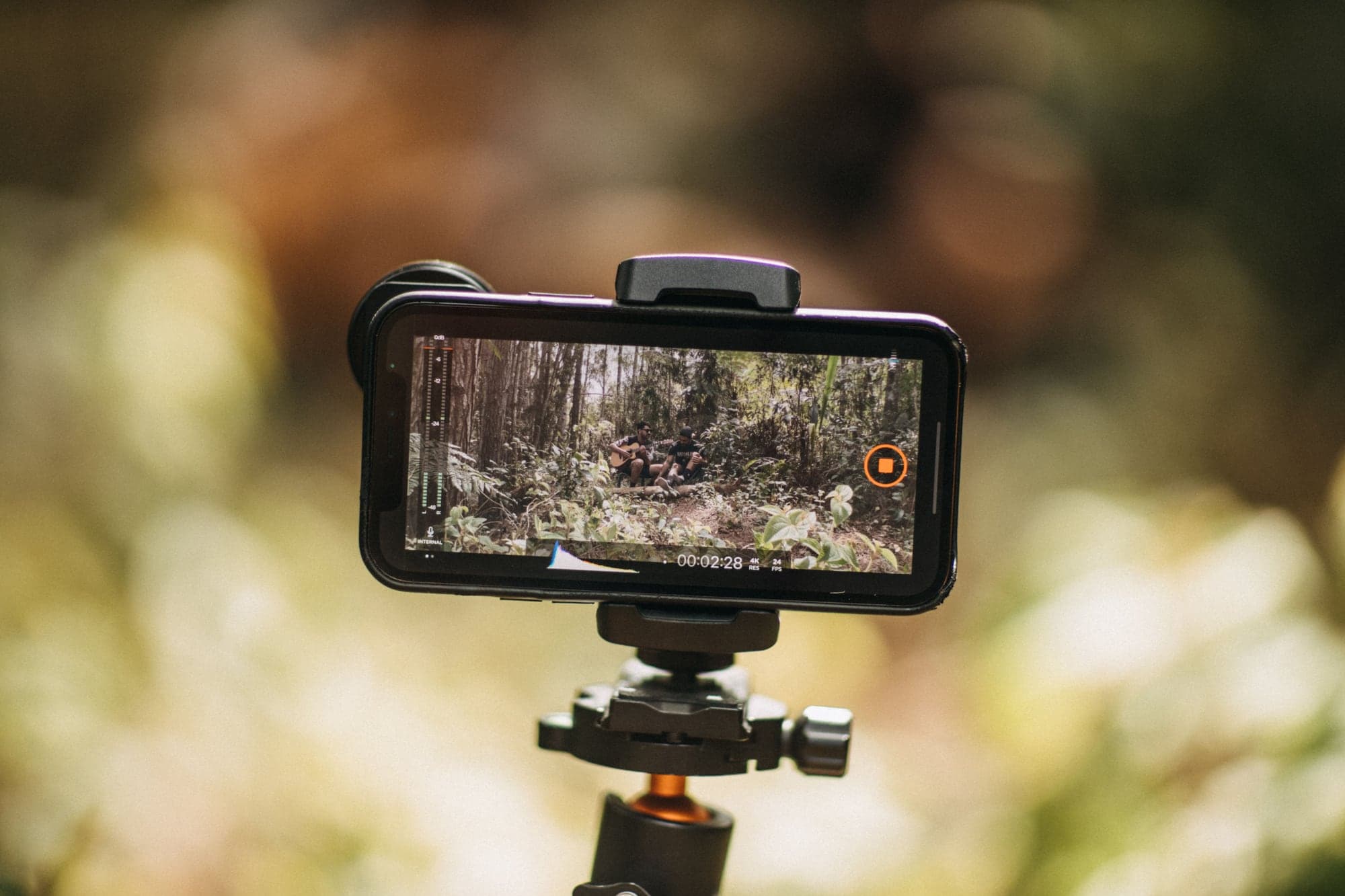 Smartphone fixé à l'horizontale sur la rotule d'un trépied, en extérieur, pendant un enregistrement : l'écran montre deux musiciens dans une forêt, un compteur à 00:02:28 et les indications 4K et 24 FPS