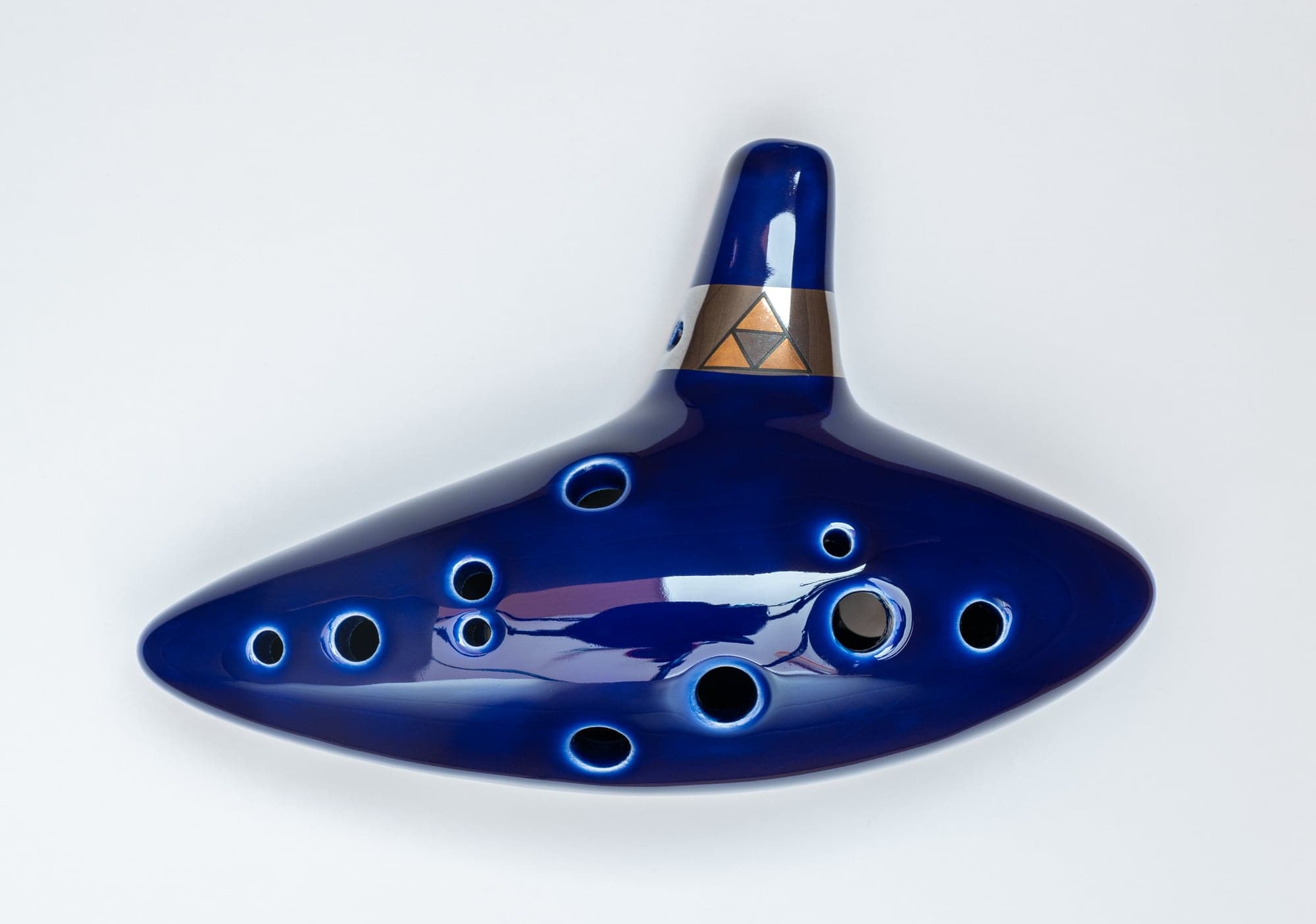 Une réplique en céramique bleue de l'ocarina du temps, l'instrument de The Legend of Zelda: Ocarina of Time, percée de trous et ornée d'une Triforce dorée sur une bague argentée, photographiée sur fond blanc.